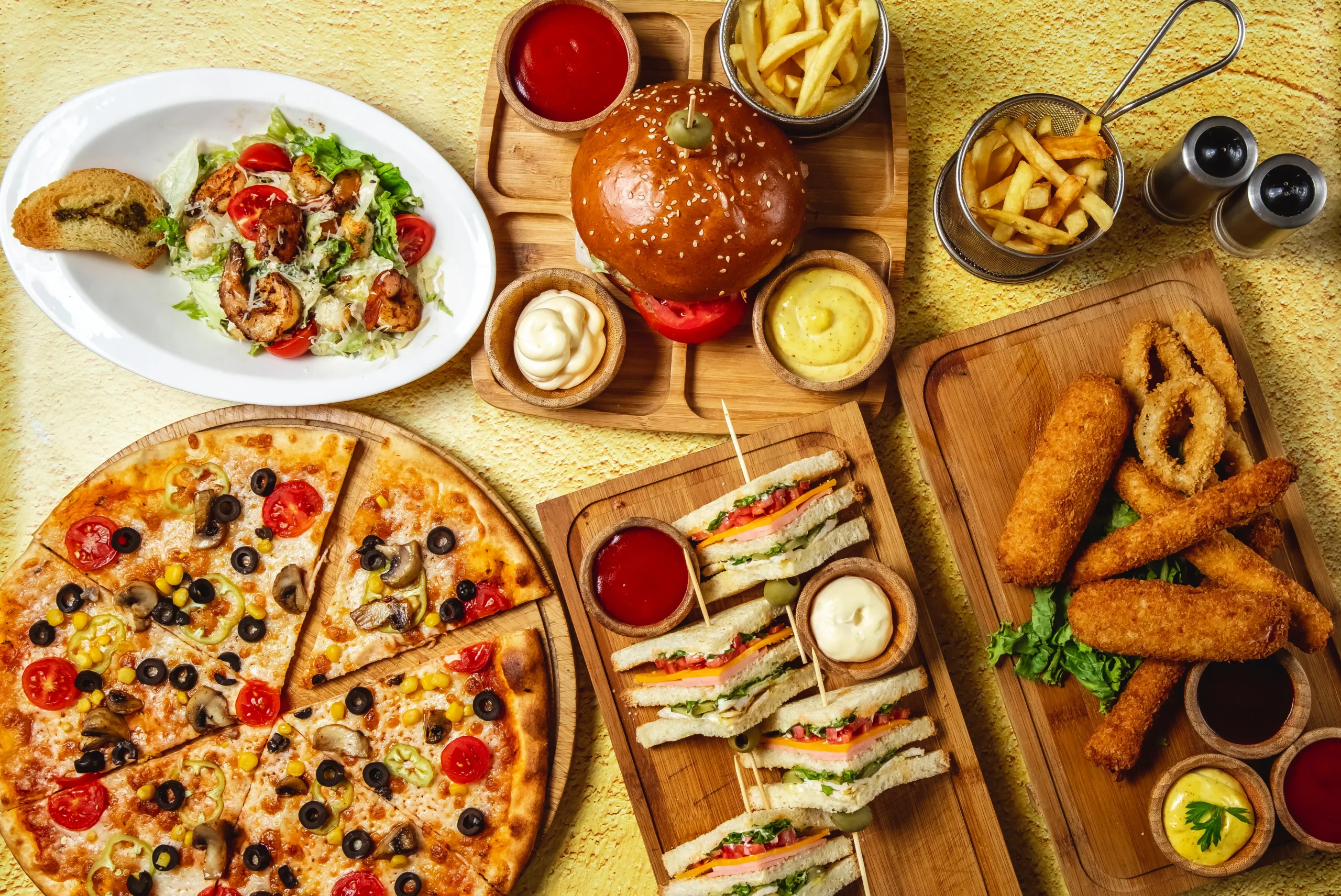 top-view-fast-food-mix-mozzarella-sticks-club-sandwich-hamburger-mushroom-pizza-caesar-shrimp-salad-french-fries-ketchup-mayo-cheese-sauces-table نبذه عن بهانس الاعاشة